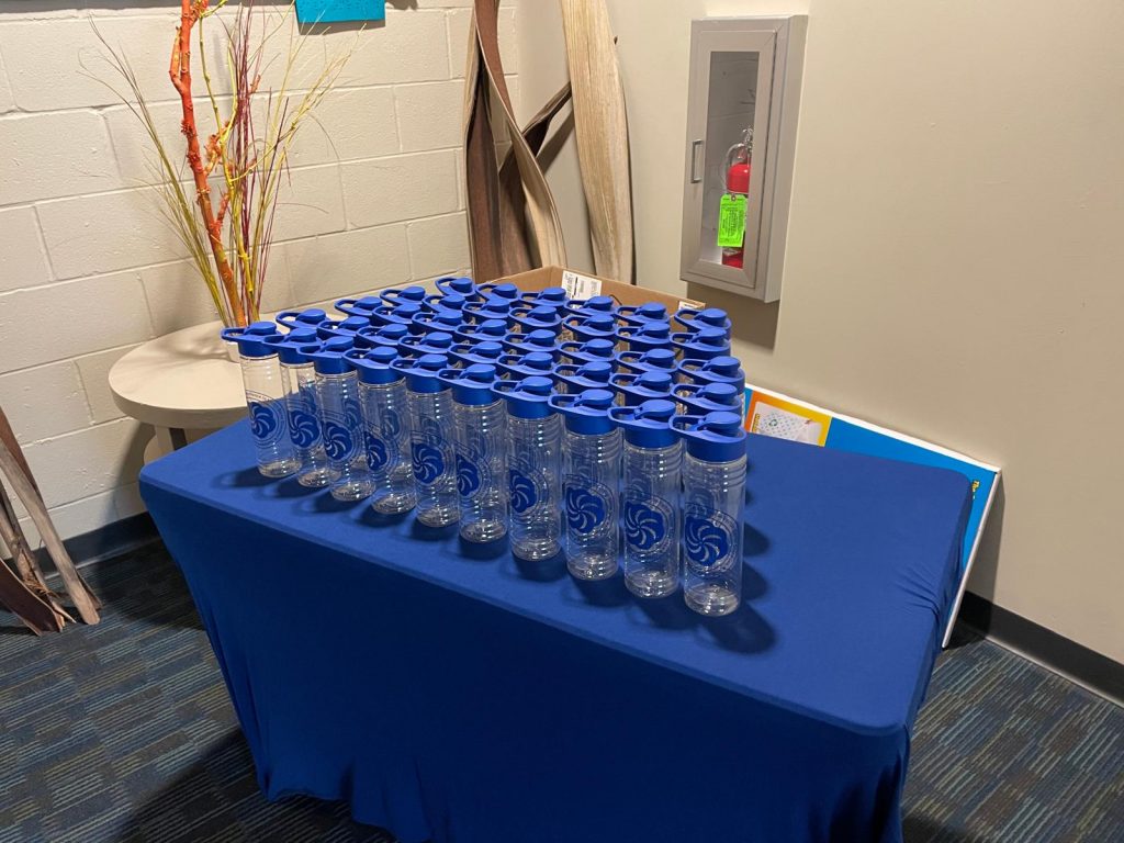 Free swagger table. A table covered with a blue cloth displays rows of clear water bottles with blue lids and the SECVBD logo
