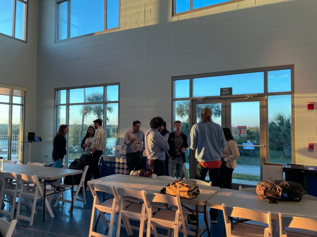 A group of SECVBD members stands and talks near large windows inside a bright room with tables and chairs set up.
