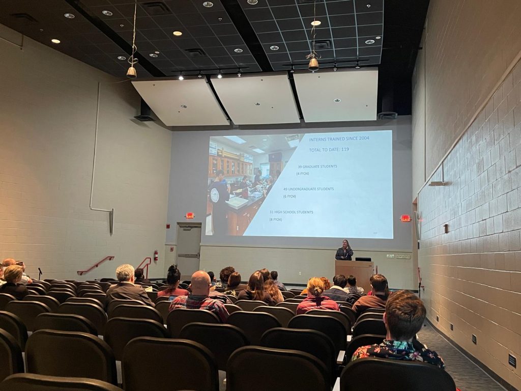 An audience sits in an auditorium watching a presenter speak at a podium in front of a large projected slide.