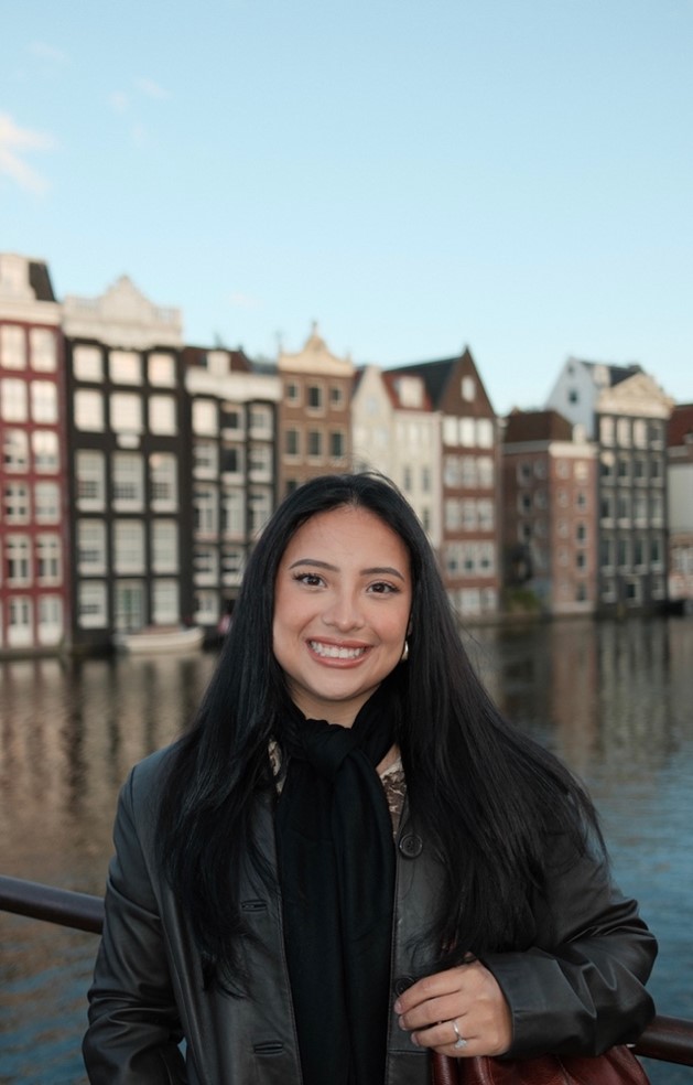 Picture of Andrea Victoria Arce, BS, standing on a bridge beside a canal with a row of tall, narrow, traditional European-style buildings lining the water in the background.