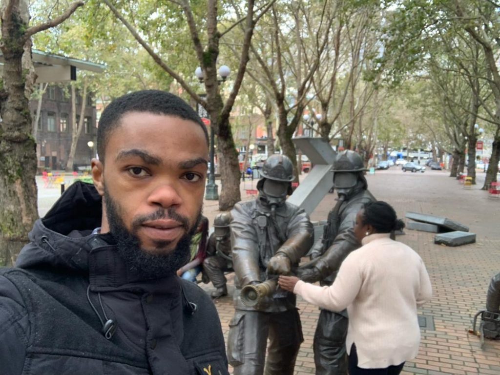Picture of Bright Agbodzi, MS MPhil, in an outdoor plaza lined with trees, while another person interacts with a group of life‑sized firefighter statues arranged as if operating a fire hose