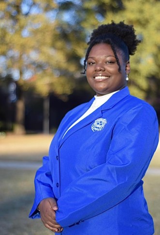 Picture of Shania Sanderson, M.S. standing outdoors in a natural, sunlit setting, wearing a bright royal‑blue blazer with an embroidered emblem on the chest and a white shirt underneath, posed with hands clasped in front against a softly blurred background of trees and grass.