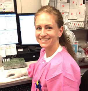 Picture of Tiffany R. Molinick, MPH, MLS(ASCP)CM wearing a bright pink lab coat and disposable gloves is seated at a workstation in a laboratory, working at a desk with multiple computer monitors and various lab supplies on shelving in the background.