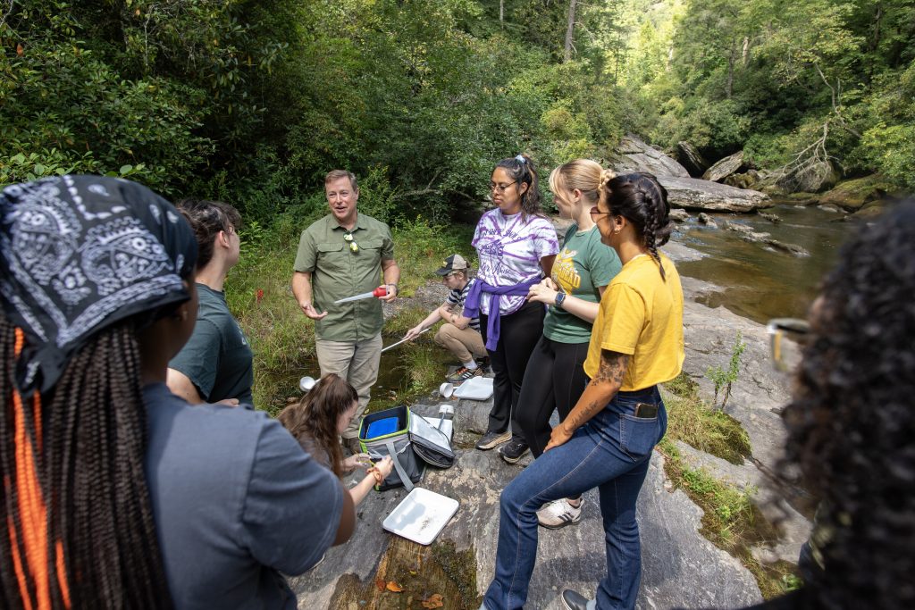 Picture of Dr Byrd and a group of trainees gathered on a rocky area beside a forested river, working together with field equipment such as trays, dippers, and basting pipettes