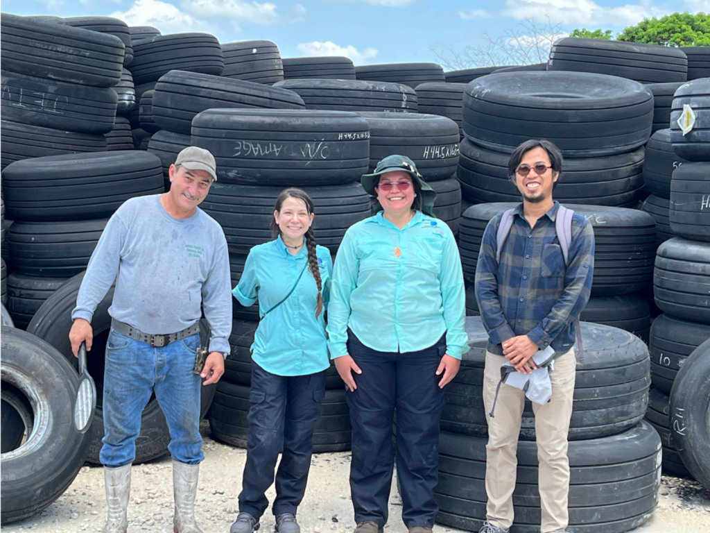 This picture shows four researchers standing side‑by‑side outdoors in front of a large stack of used vehicle tires. The tires are piled high behind them, forming several tall rows that extend across the background.